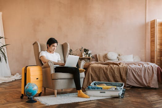 A man uses a laptop while packing a suitcase at home, ready for travel.