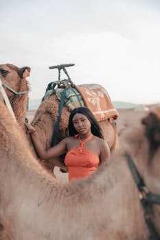 A young woman in a red dress stands beside camels in Marrakech, Morocco.