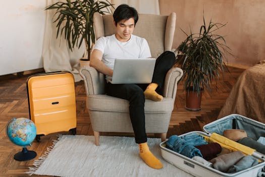 Man sitting on armchair planning travel, using laptop, with suitcases and globe nearby.