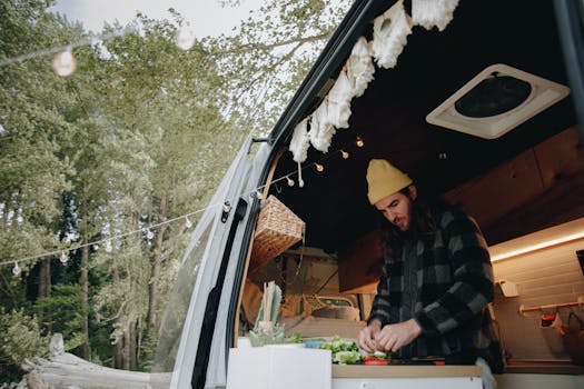 A man in a camper van kitchen prepares food amidst a scenic forest setting, showcasing van life and outdoor adventure.