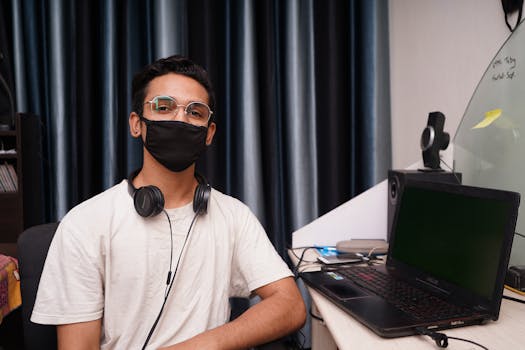 A young man wearing a mask works at a home office with a laptop during the pandemic.